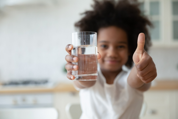 Biracial girl holding glass of water, showing thumbs up gesture.